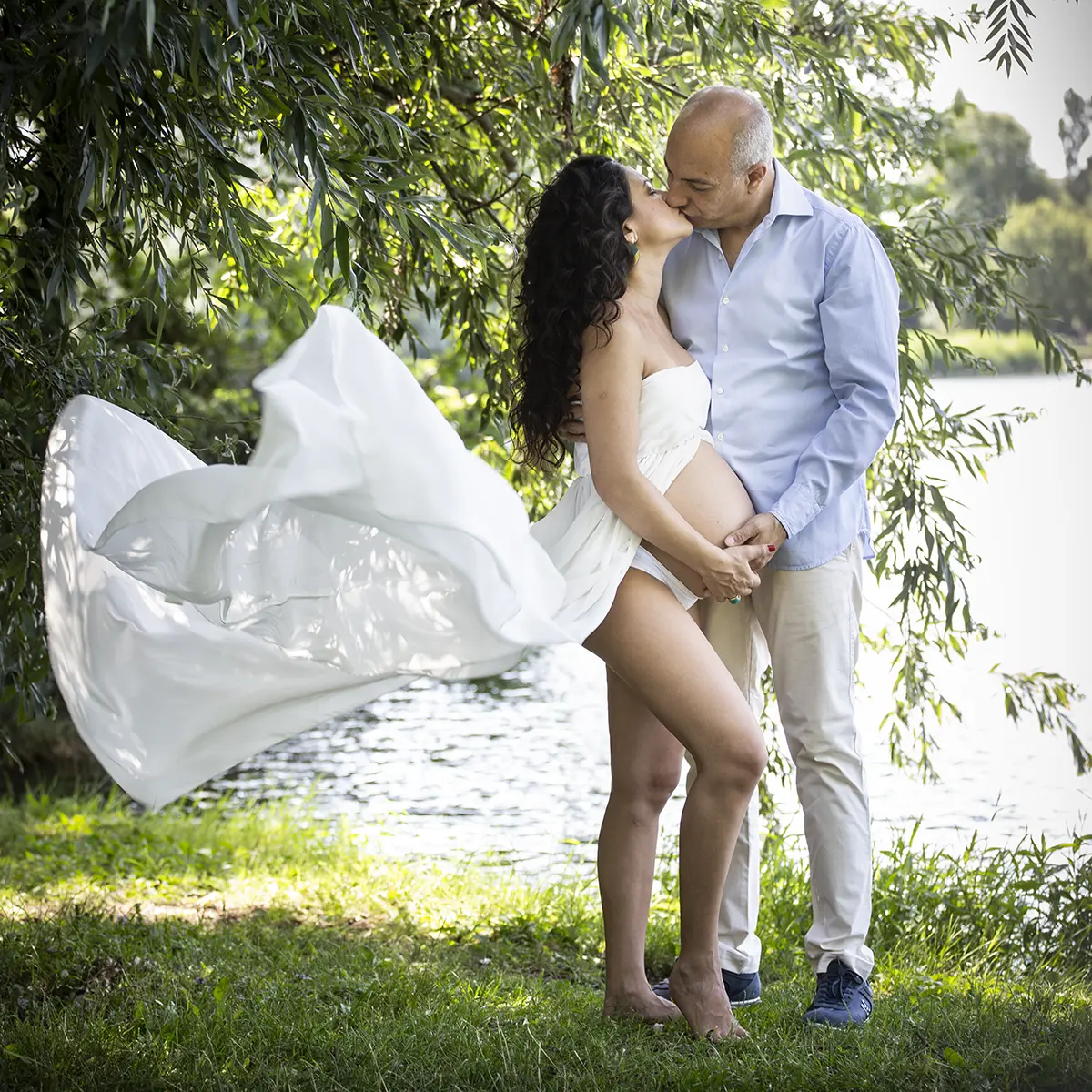 Coppia in attesa che si bacia durante un book fotografico gravidanza all'aperto a Milano, abito bianco fluttuante vicino al lago